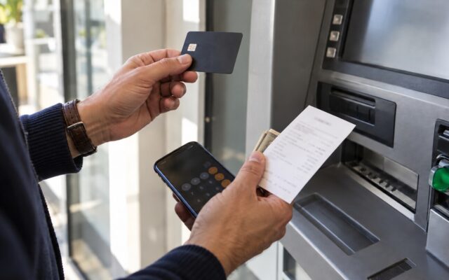Persona revisa el comprobante de un retiro frente a un cajero, con tarjeta genérica y calculadora en el teléfono.