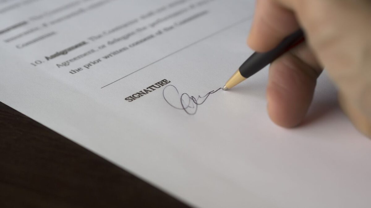 Una mano firma un contrato sobre un escritorio de madera